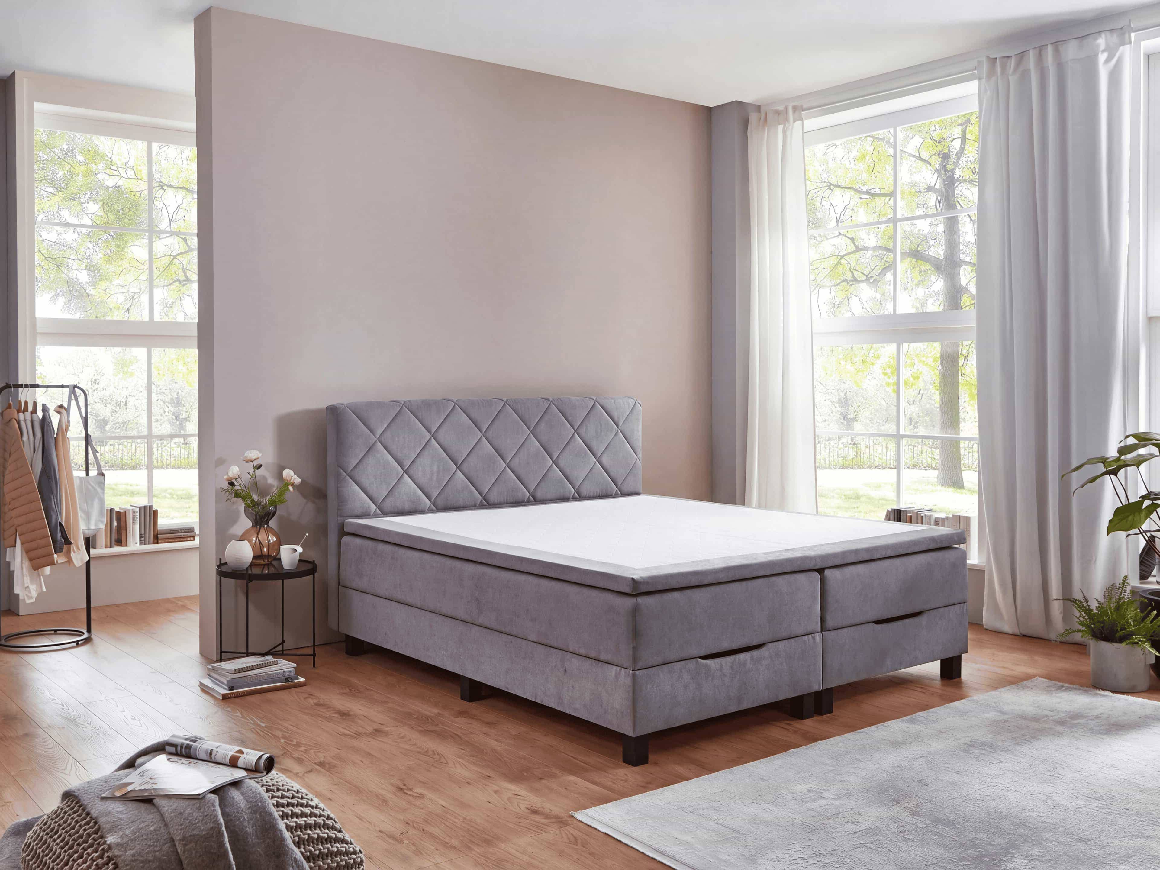 Boxspringbett mit grauem Stoffbezug, gestepptem Kopfteil und schwarzen Bettfüßen in L-Form.