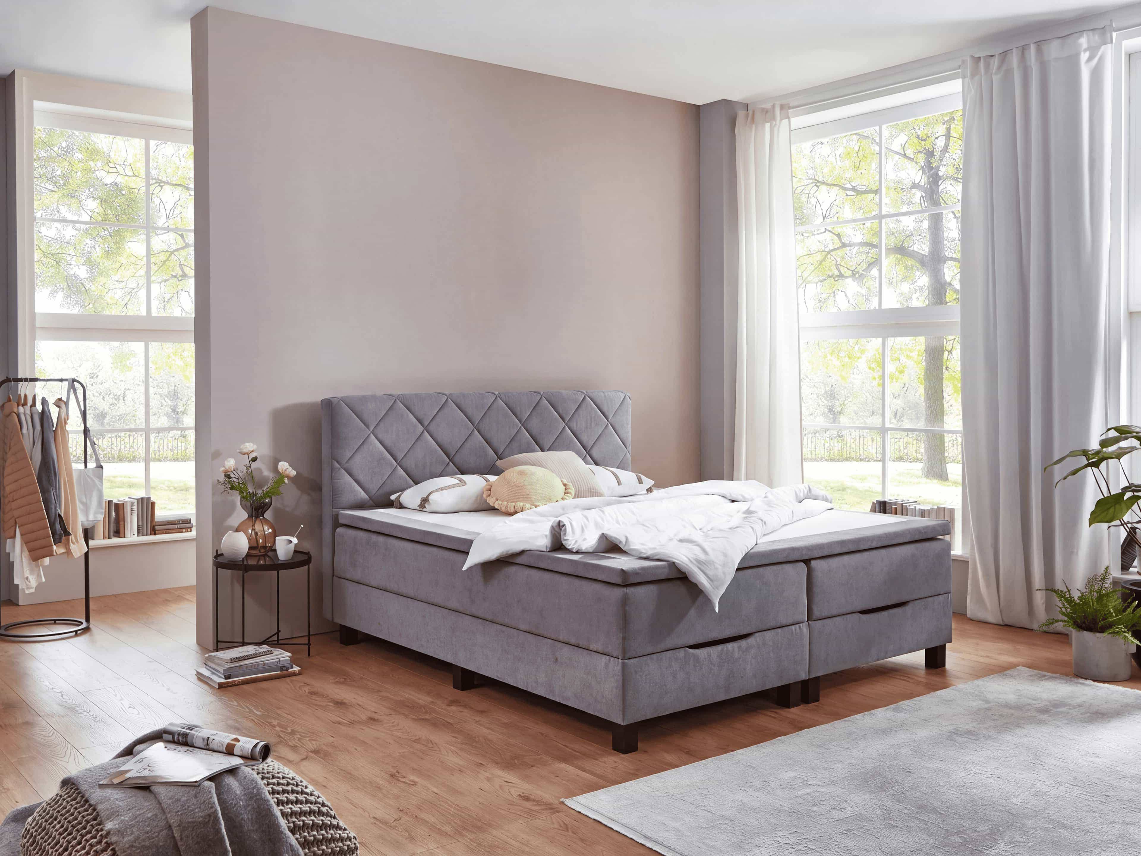 Boxspringbett Kabs Modell in Hellgrau mit gestepptem, gepolstertem Kopfteil und dunklen Füßen