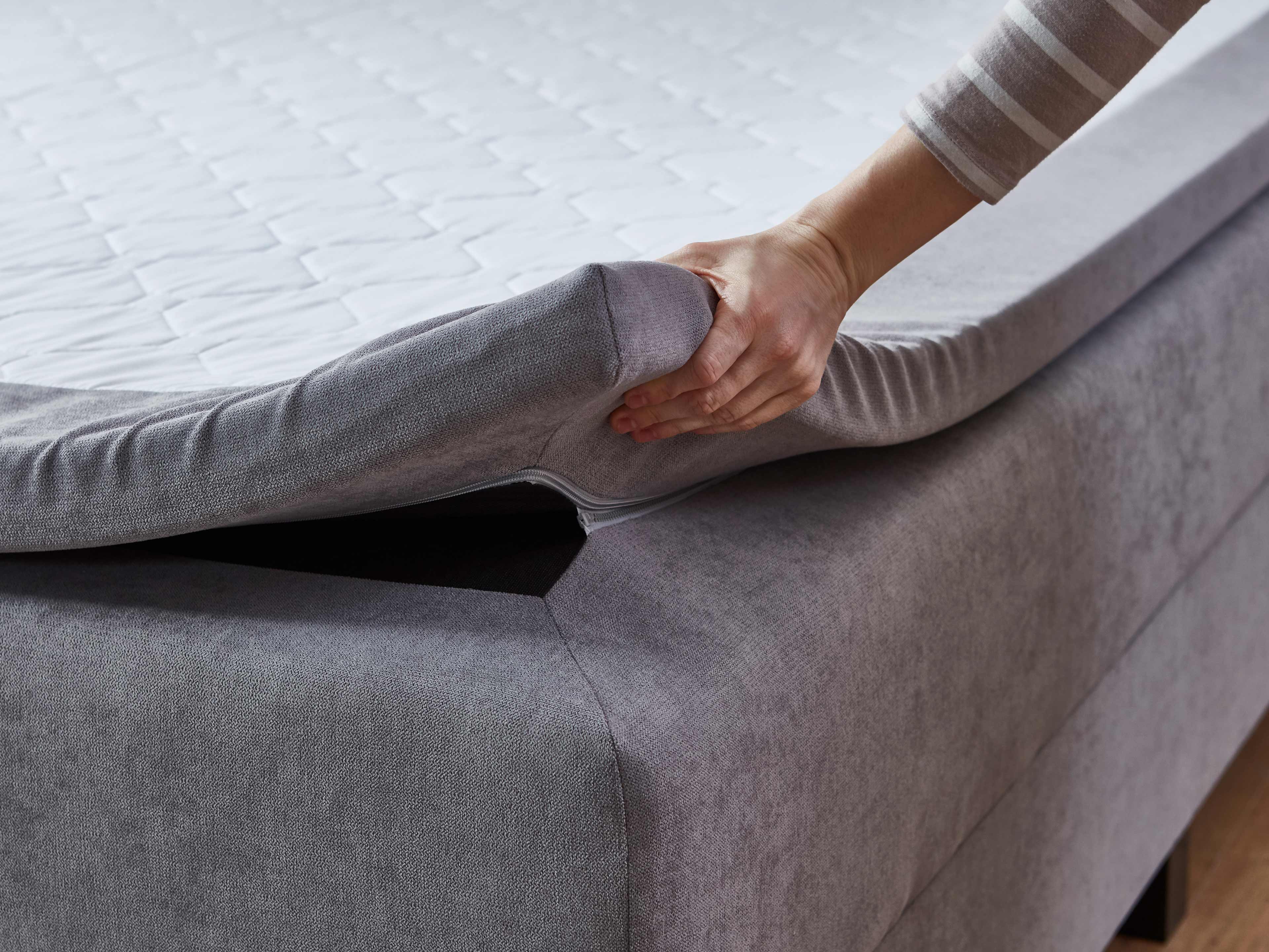 Boxspringbett mit grauem Stoffbezug, seitliche Ansicht, abnehmbare Matratzenauflage, Hand hält Bezug