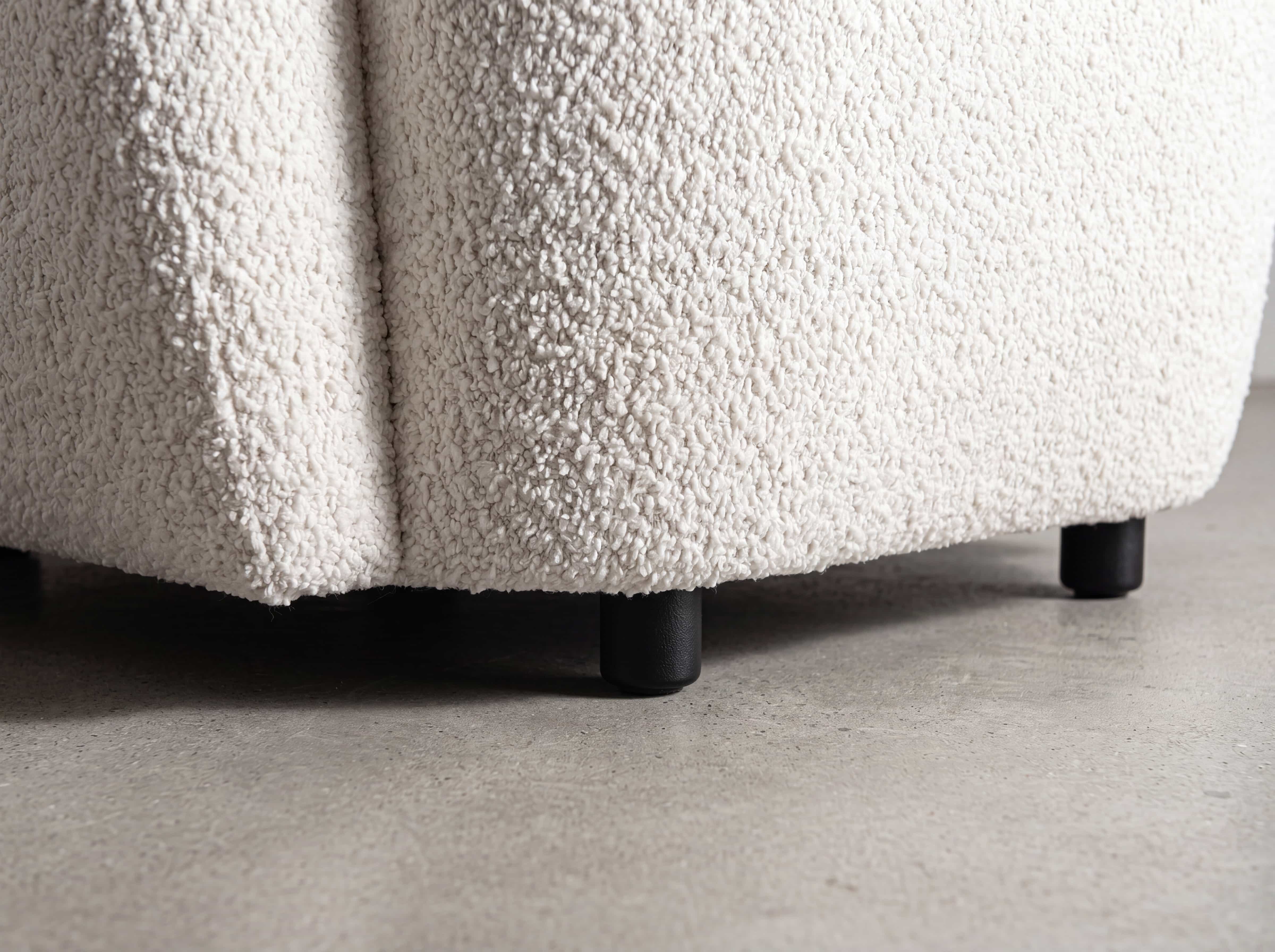 Nahaufnahme eines Sofafußes mit cremefarbenem, strukturiertem Stoffbezug und schwarzen runden Füßen.