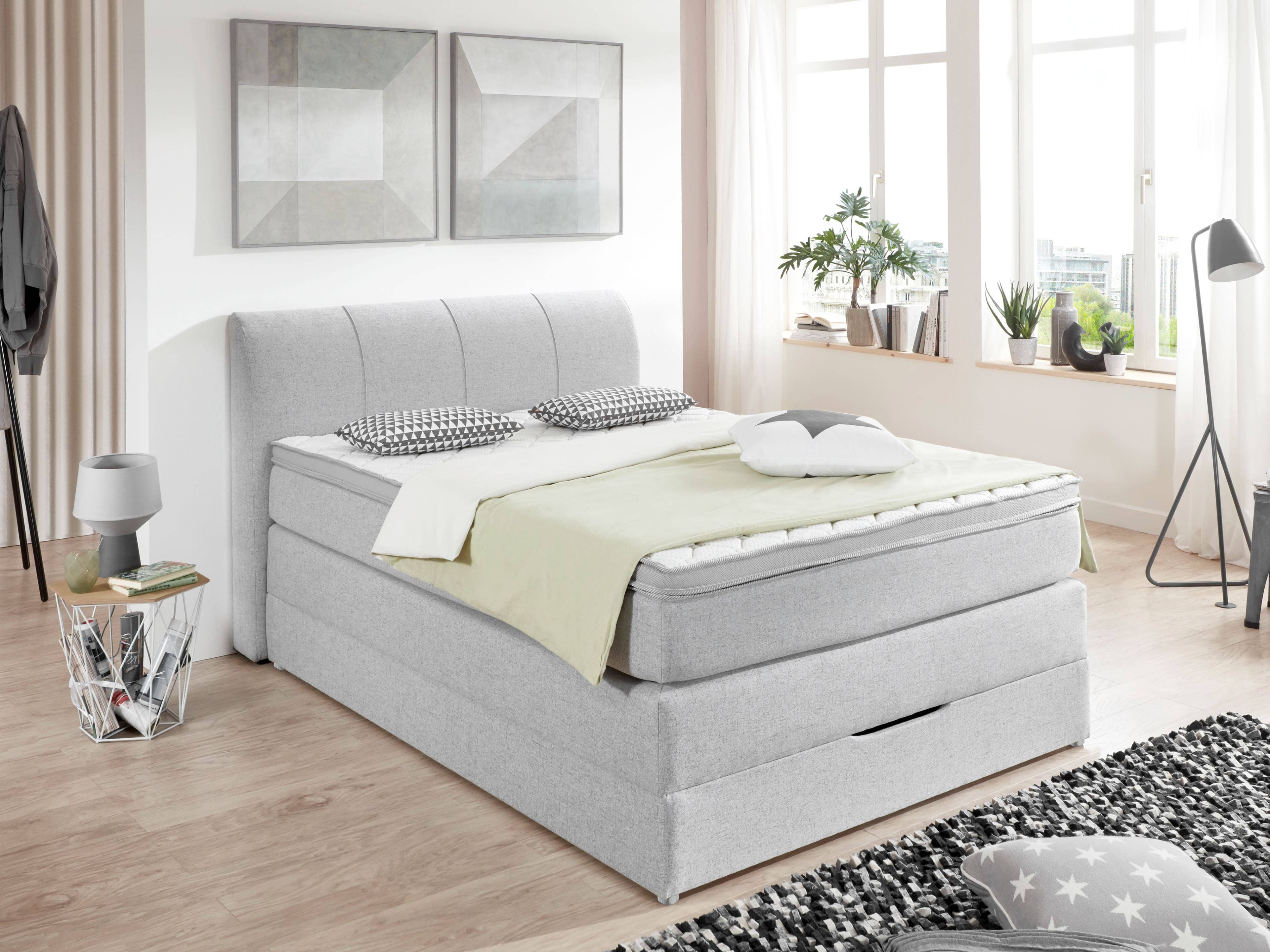 Boxspringbett mit hellgrauem Stoffbezug, gepolstertem Kopfteil und sichtbarem Stauraum-Fach