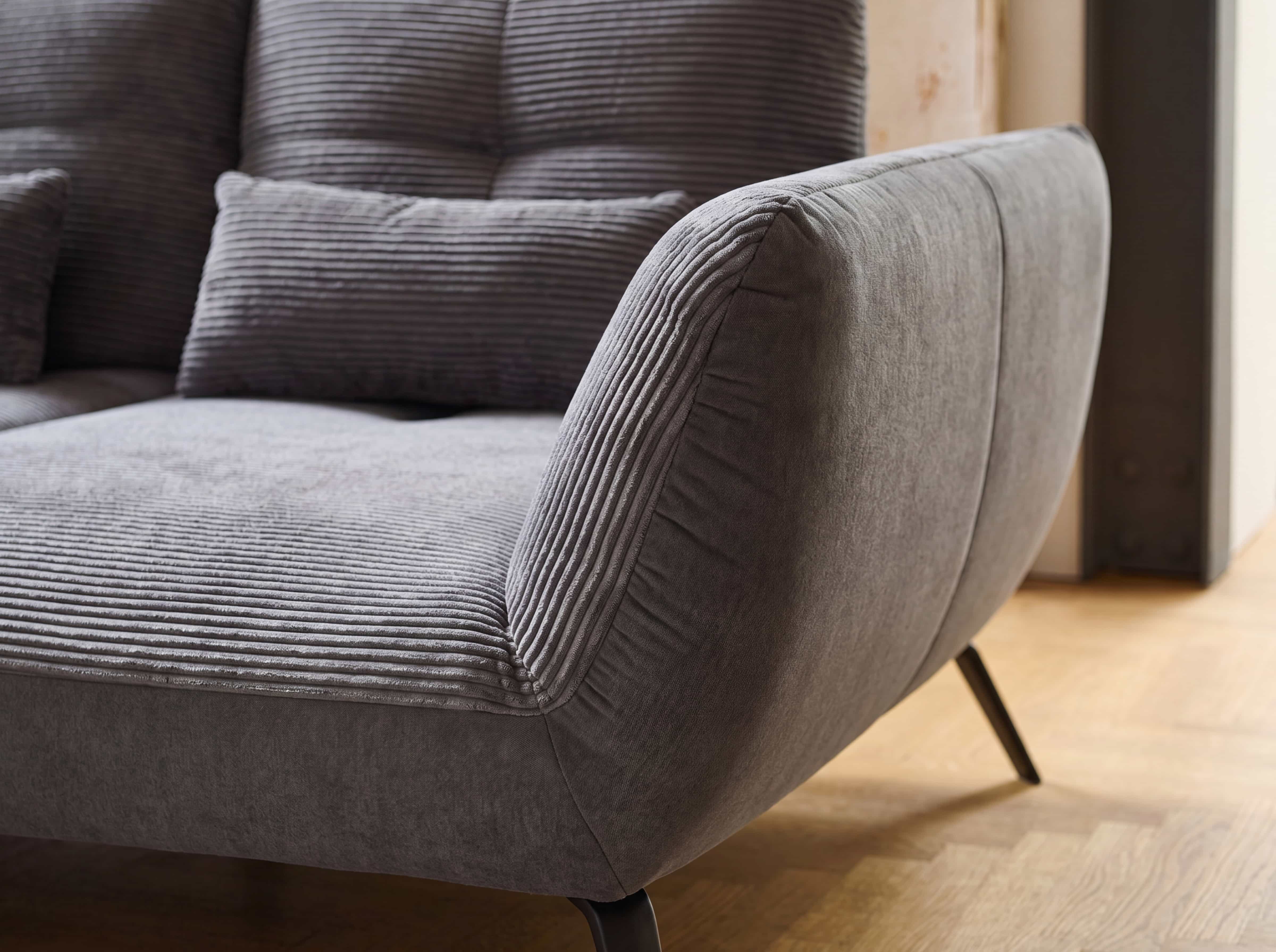 Ecksofa Paola – Detailansicht mit grauem Cordbezug, schräger Armlehne und Zierkissen
