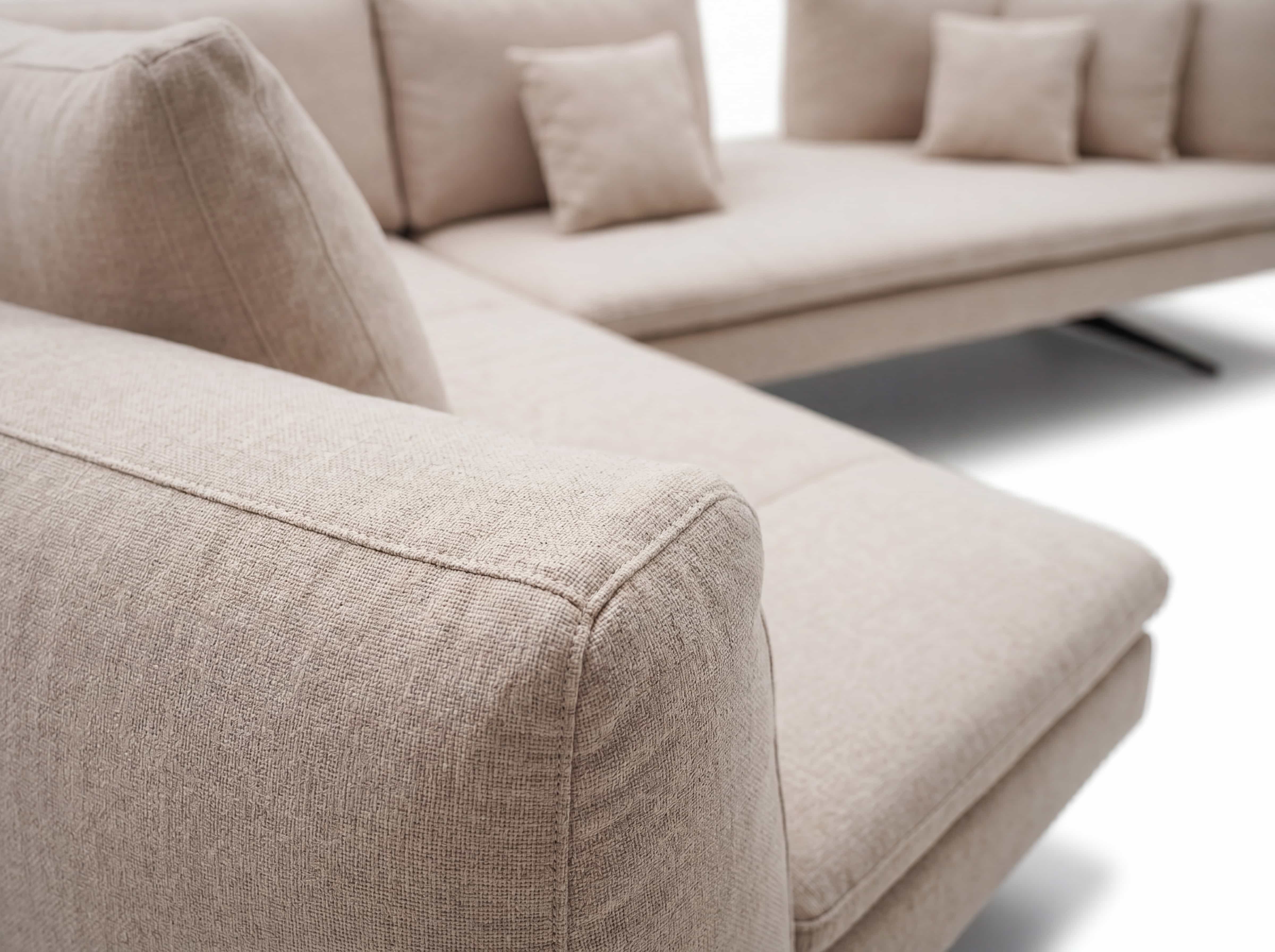 Ecksofa mit beigefarbenem Stoffbezug, gepolsterter Sitzfläche, Armlehnen und losen Rückenkissen