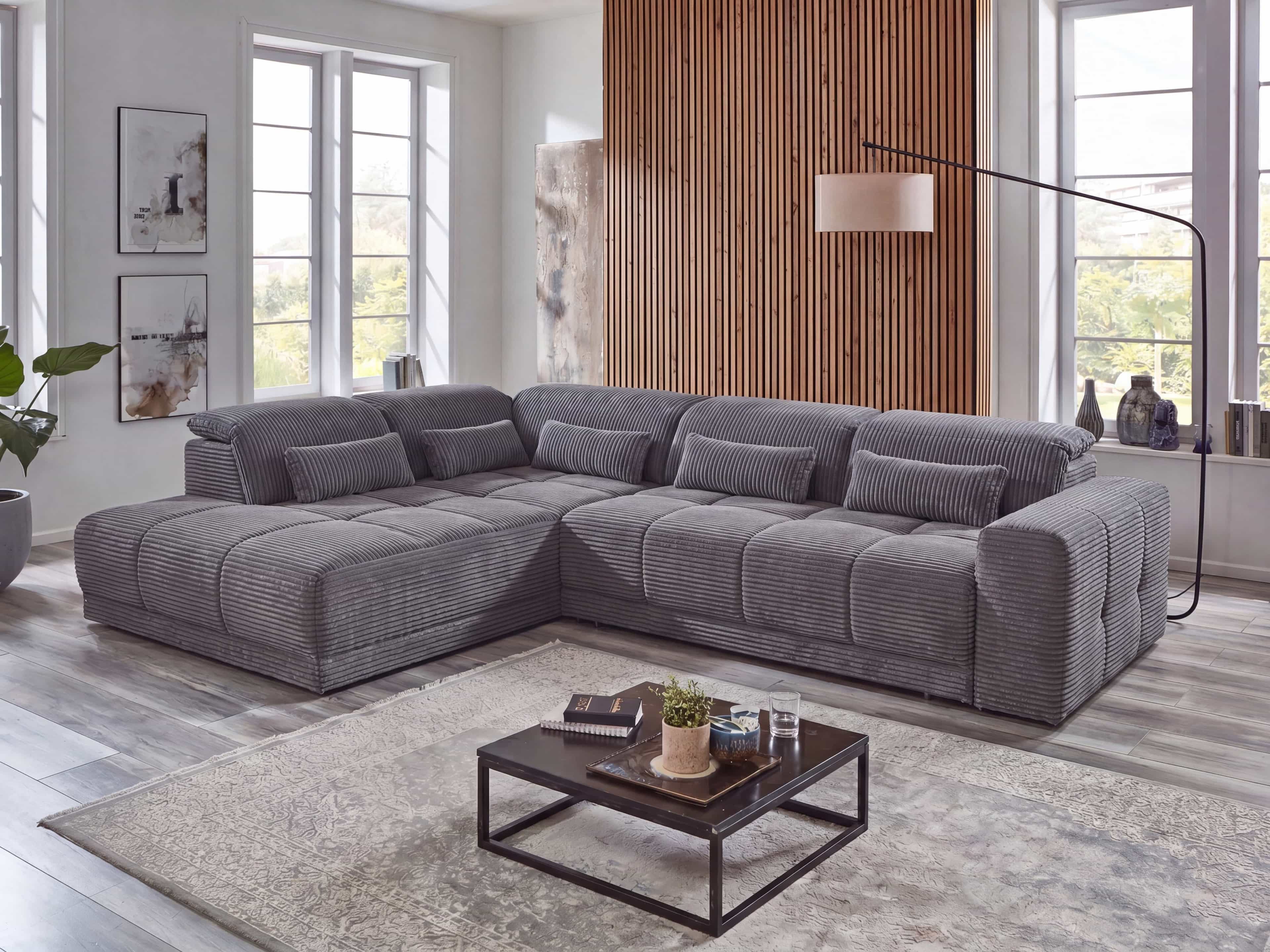 Ecksofa mit grauem Cordbezug, breiten Armlehnen und mehreren Rücken- und Zierkissen