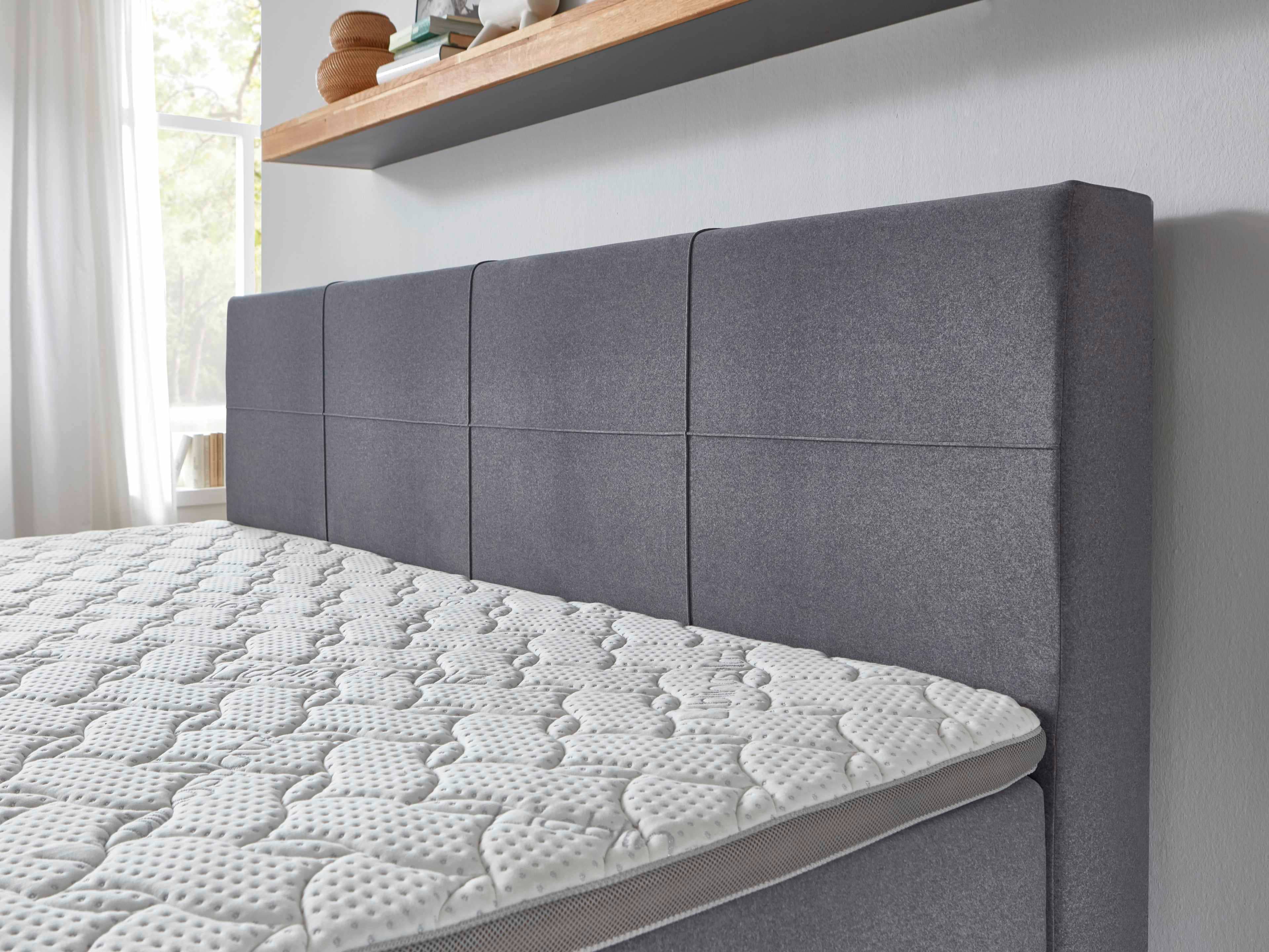 Boxspringbett mit grauem, gepolstertem Kopfteil aus Stoff und sichtbarer, gemusterter Matratze