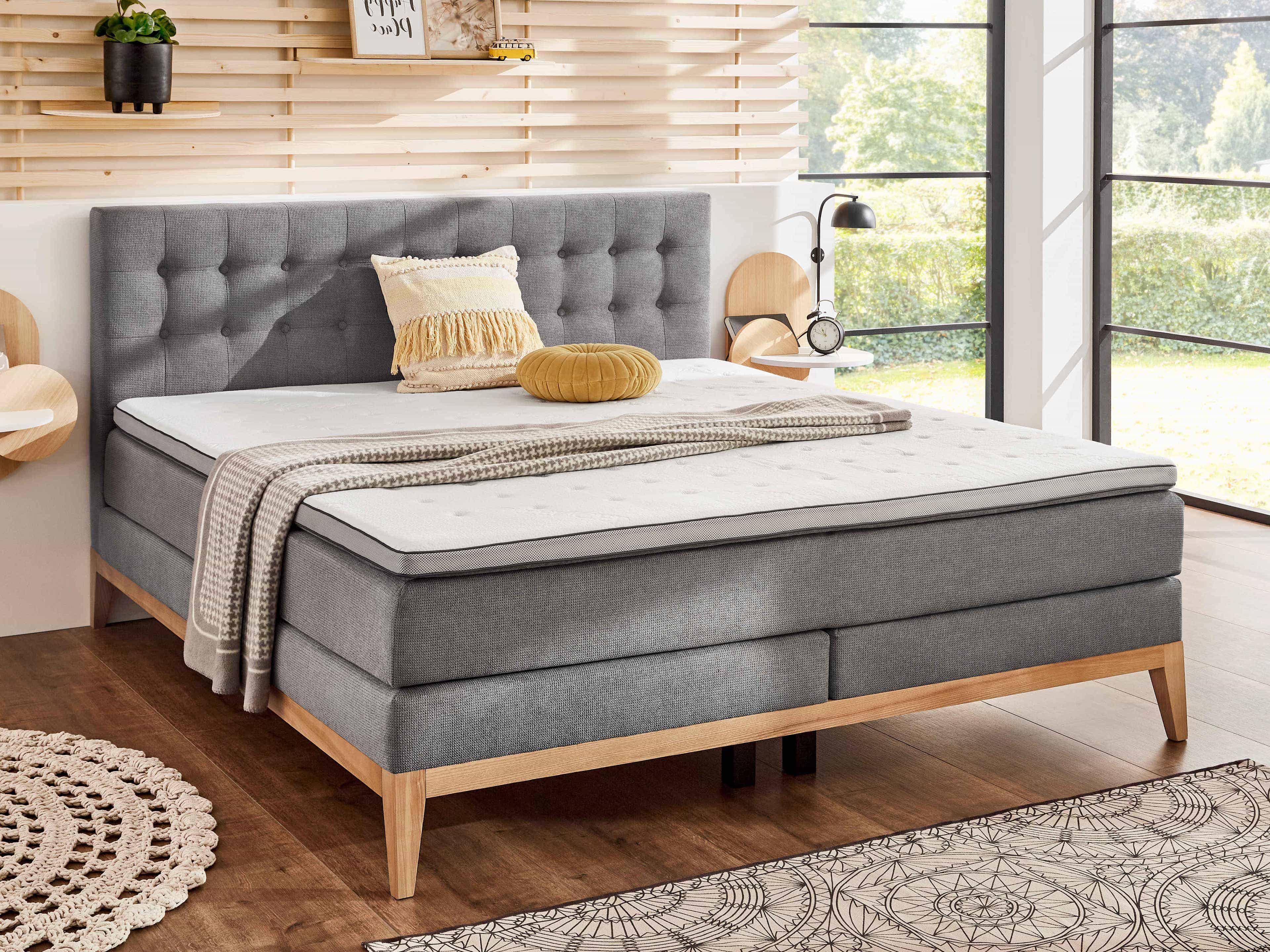 Boxspringbett mit grauem Stoffbezug, gestepptem Kopfteil und Holzfüßen, rechteckige Form