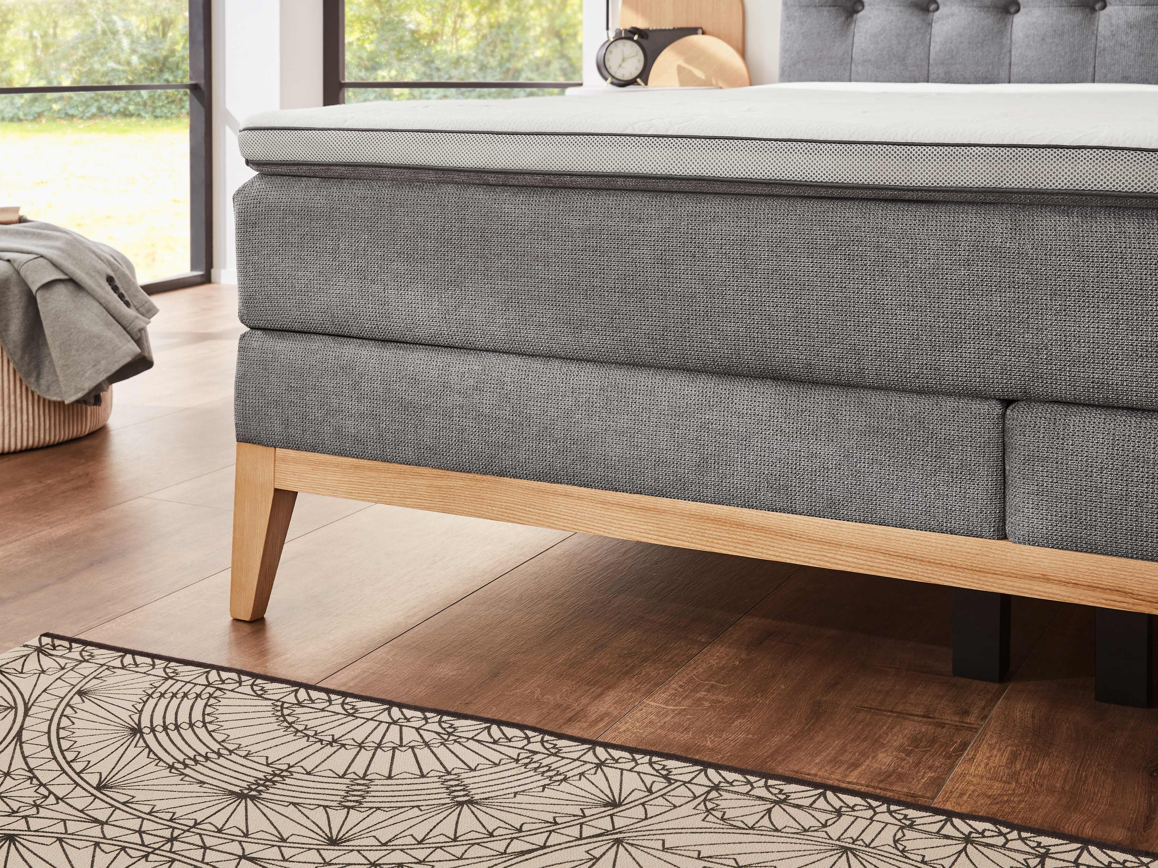 Boxspringbett mit grauem Stoffbezug, hellem Holzrahmen, sichtbarem Kopfteil und gepolsterter Liegefläche