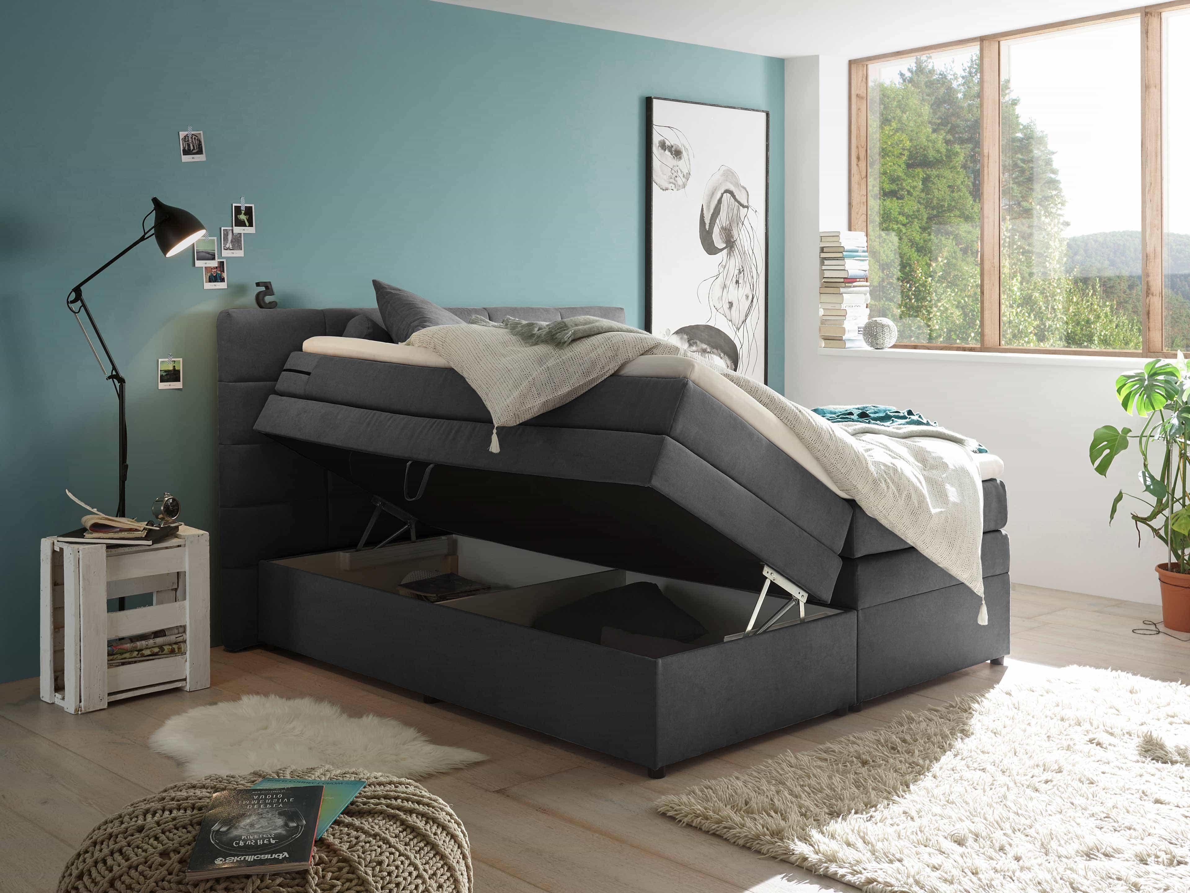Boxspringbett mit grauem Stoffbezug, gepolstertem Kopfteil und aufgeklappter Bettkastenfunktion