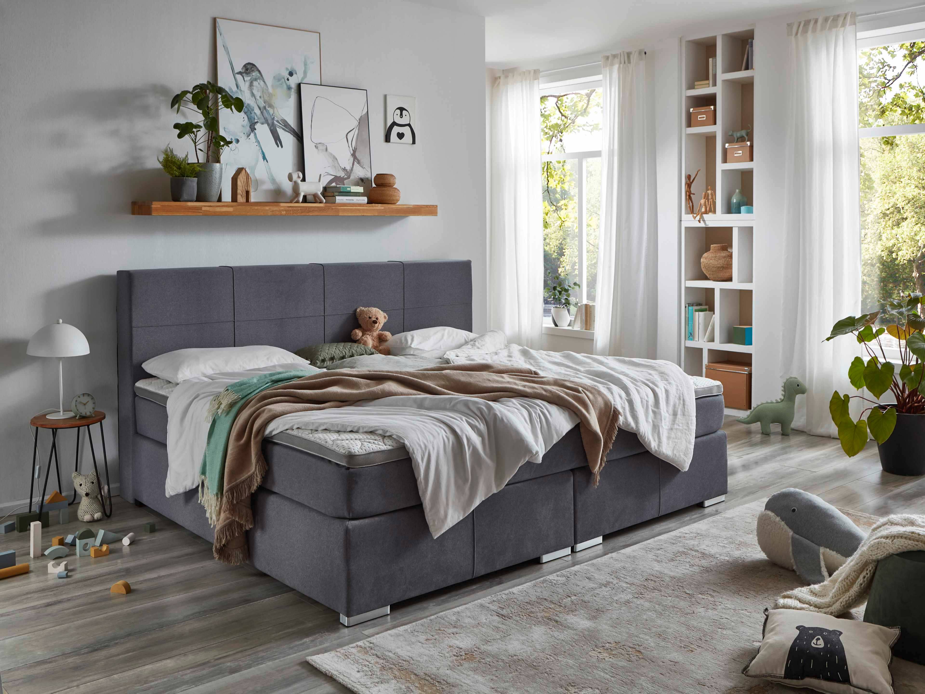 Boxspringbett mit grauem Stoffbezug, gepolstertem Kopfteil und sichtbaren Metallfüßen