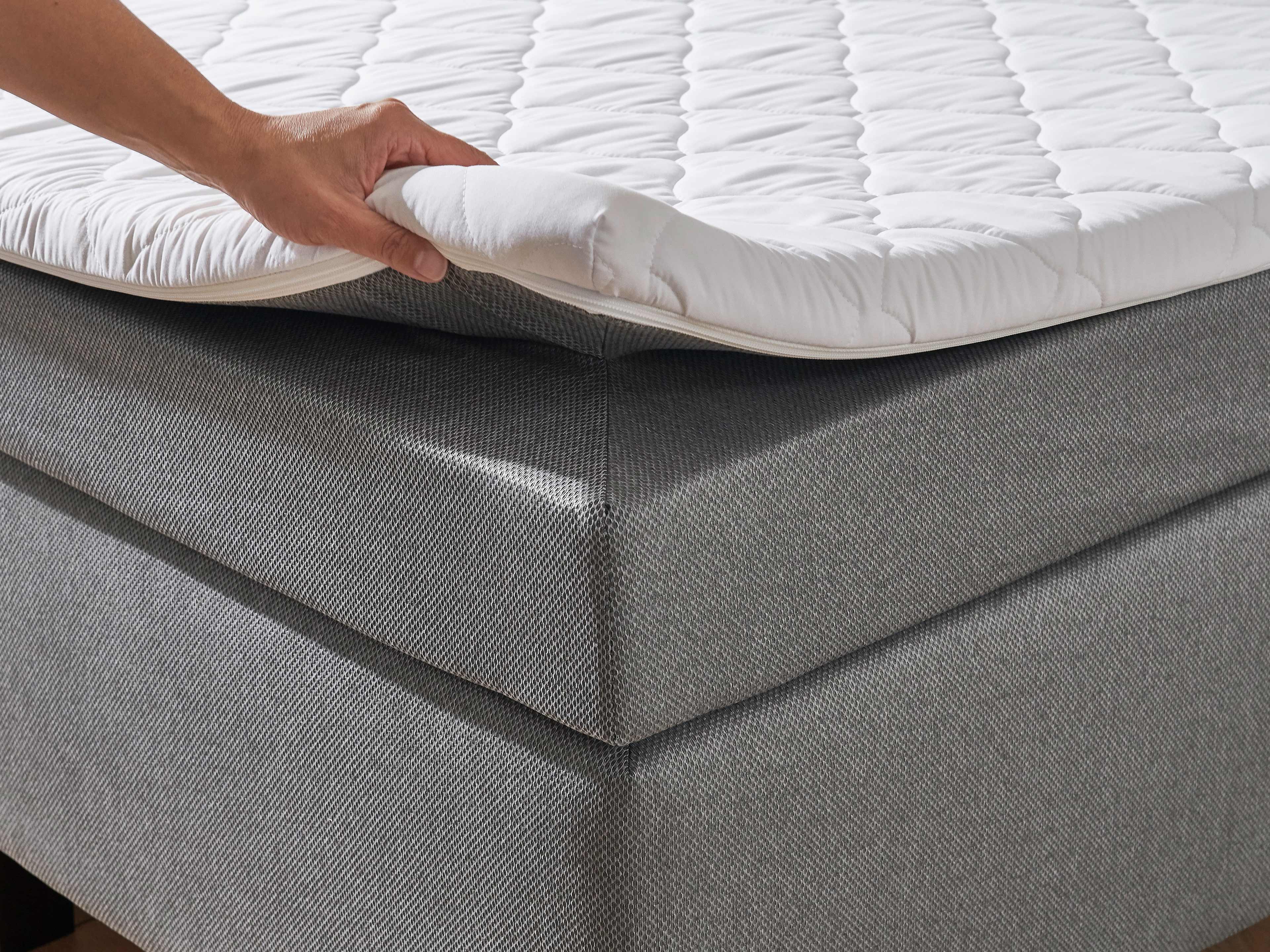 Boxspringbett mit grauem Stoffbezug aus Stoff, sichtbarer weiß gesteppter Topper, Eckansicht